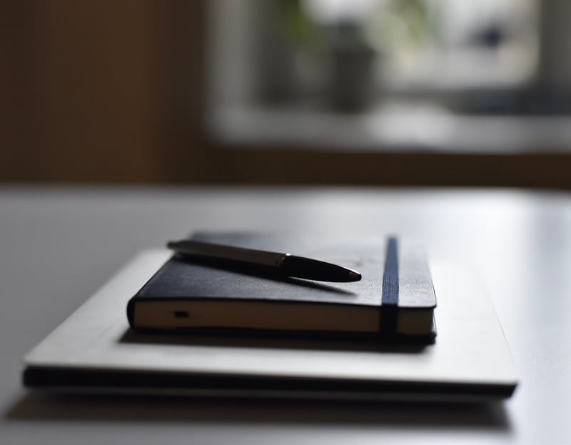 En silverfärgad bärbar dator, en blå anteckningsbok och en penna ligger på ett bord. Ett fönster finns i bakgrunden.